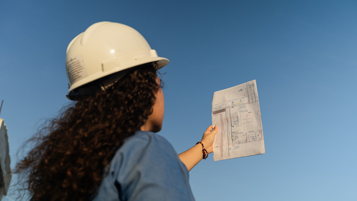 Ingeniero revisa planos arquitectónicos con cielo azul de fondo.